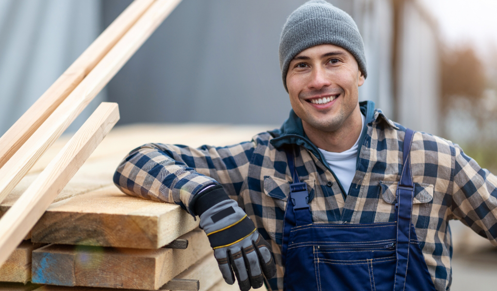 man in flannel shirt, overalls, and beanie next to wood pieces
