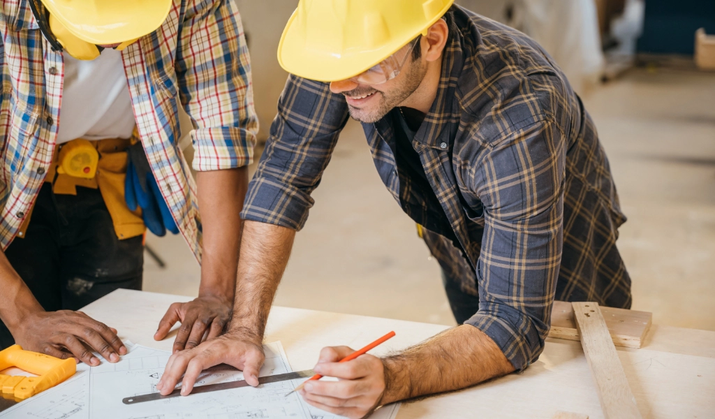 Two men in yellow hard hats work on blueprints together.